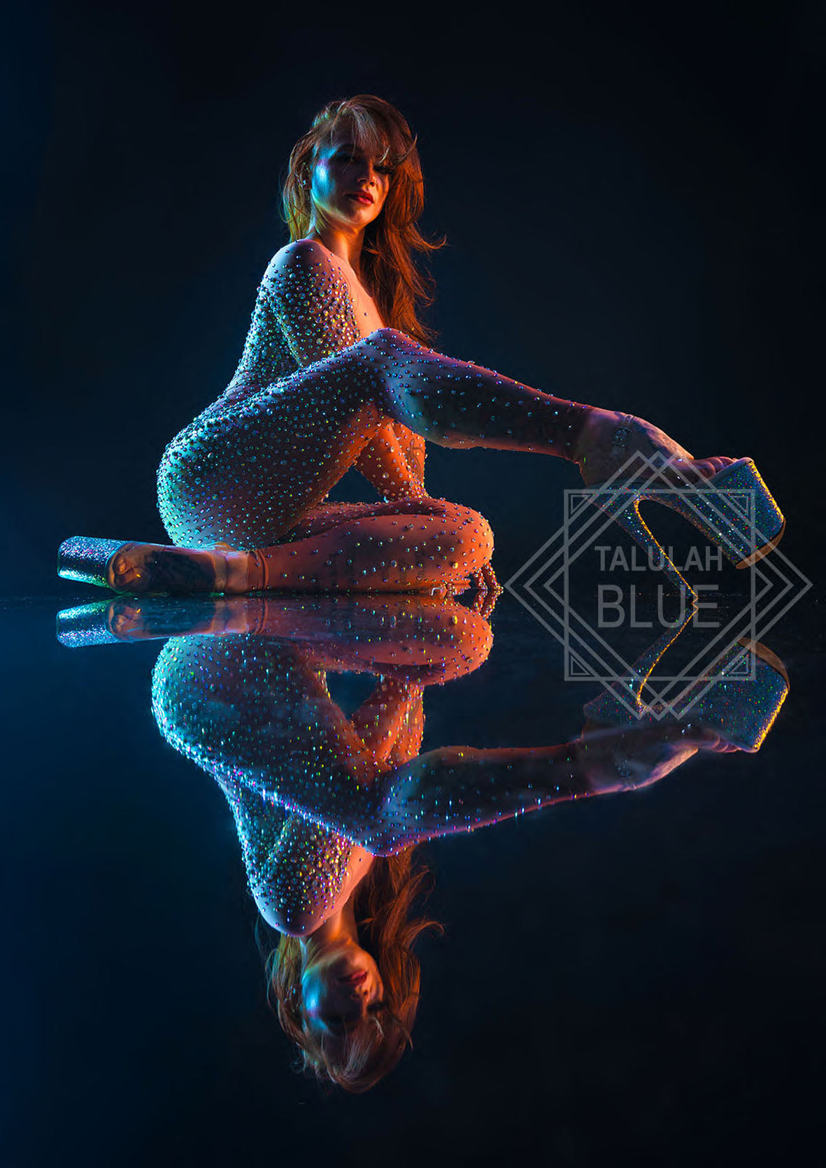 Person wearing a sparkling outfit sitting on a reflective surface with a dark background. Talulah Blue Costumes by Andre Pattenden